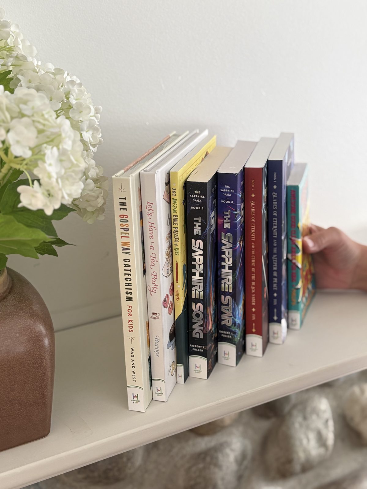 A row of Harvest Kids books standing spine-out on a white shelf beside white hydrangeas
